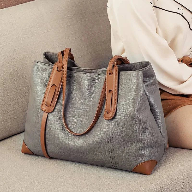 Bolsa de ombro feminina em couro legítimo, na cor cinza com detalhes e alças marrons, design elegante sobre sofá bege.
