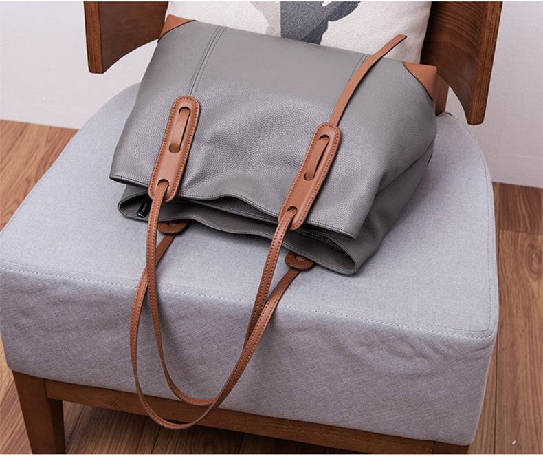 Bolsa de ombro feminina em couro legítimo cinza com alças marrons e design estruturado, vista de cima sobre cadeira estofada.