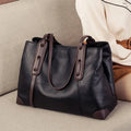Bolsa de ombro feminina em couro legítimo, na cor preta com alças e detalhes marrons, estilo sofisticado sobre sofá bege.
