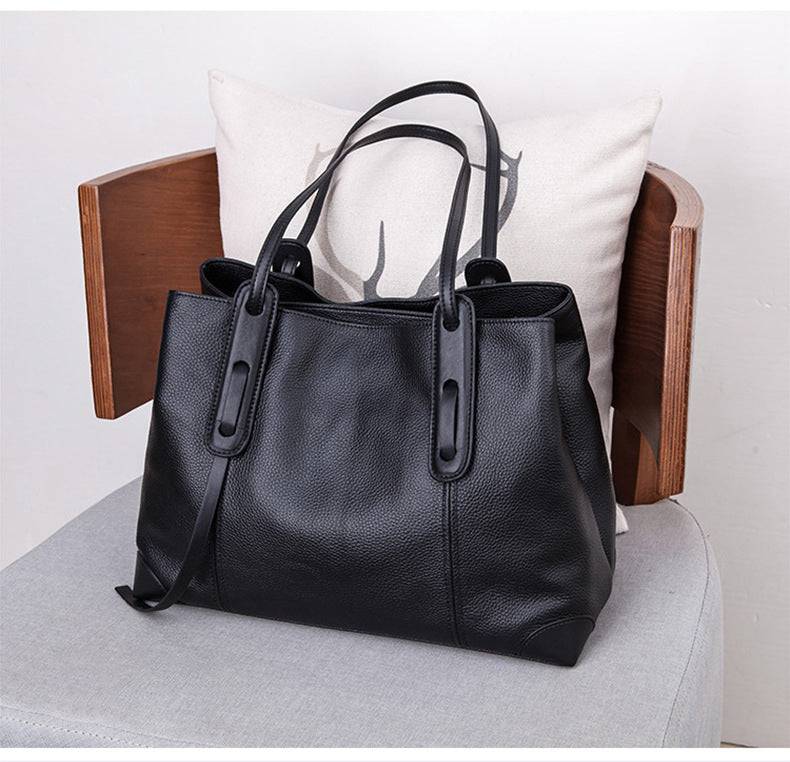 Bolsa de ombro feminina em couro legítimo, cor preta, sobre cadeira com almofada decorativa de cervo.