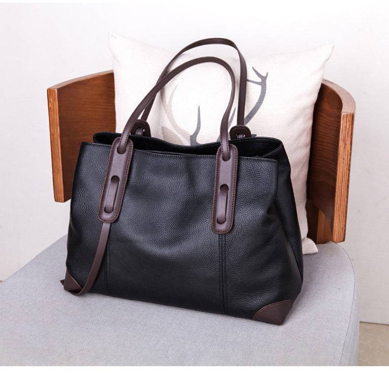 Bolsa de ombro feminina em couro legítimo, cor preta com alças e detalhes em marrom, sobre cadeira com almofada de cervo.