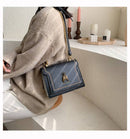 Bolsa tiracolo feminina de couro estruturada preta com alça de corrente dourada e detalhe de abelha dourada, sobreposta em ambiente minimalista com velas e objetos decorativos.