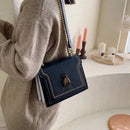 Bolsa tiracolo feminina de couro estruturada preta, alça de corrente dourada, detalhe decorativo de abelha em metal, usada por modelo vestindo suéter bege em ambiente minimalista.