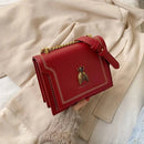 Bolsa tiracolo feminina de couro estruturada vermelha, com alça de corrente dourada e detalhe decorativo de abelha em metal, posicionada sobre casaco bege claro.