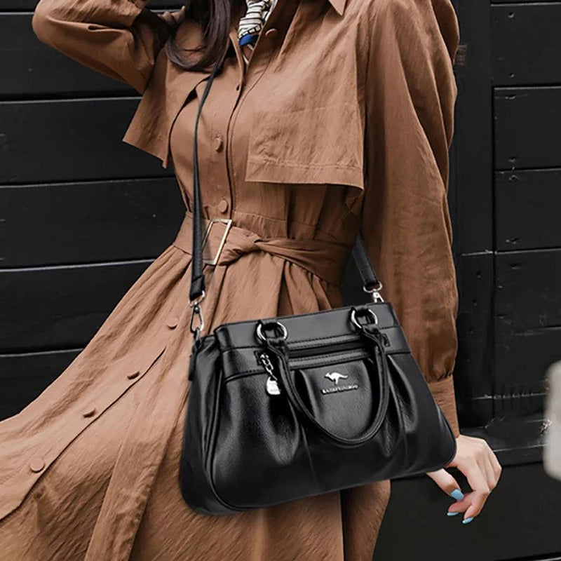 Bolsa Tote Estruturada Feminina Couro Radiance preta. Alças de mão, alça transversal, zíper frontal com charm, logo metálico. Usada por modelo em trench coat.