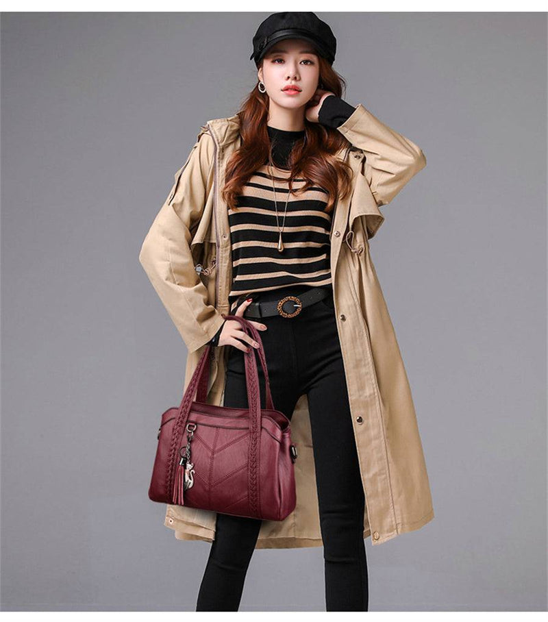 Mulher segurando bolsa tote estruturada vinho com alças trançadas, combinando com trench coat bege, blusa listrada e calça preta, em look de outono.