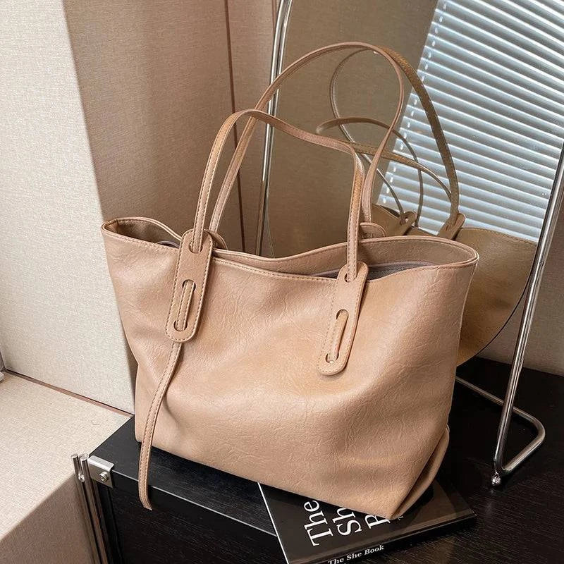 Bolsa tote feminina em couro bege claro, com design minimalista e alças longas, apoiada em mesa com livros
