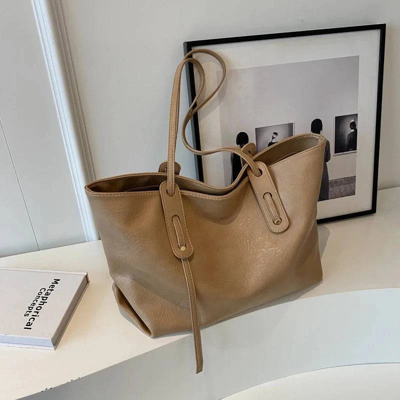 Bolsa tote feminina em couro bege, com design minimalista, detalhes de costura e alças finas, posicionada sobre mesa branca com livros e quadros