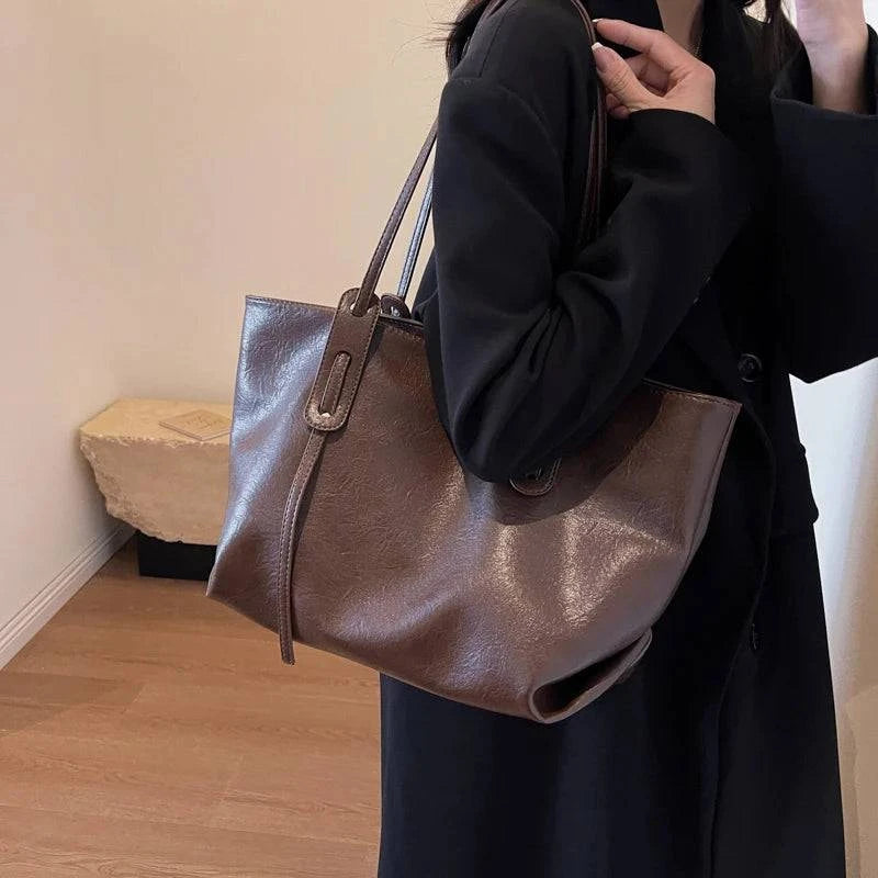 Bolsa tote feminina de couro marrom escuro com acabamento liso, alças longas e design minimalista, usada no ombro com blazer preto