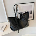 Bolsa tote feminina em couro preto, design elegante com alças finas e detalhes de costura, exibida sobre mesa branca com livros e quadros minimalistas