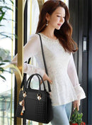 Mulher com blusa de renda branca e calça clara usando bolsa tote estruturada preta com alça transversal, exibindo detalhes texturizados e chaveiro com borlas.