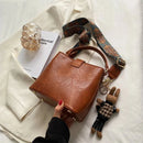 Bolsa tote transversal de couro marrom com alça estampada e bolso frontal, acessórios e chaveiro de coelho