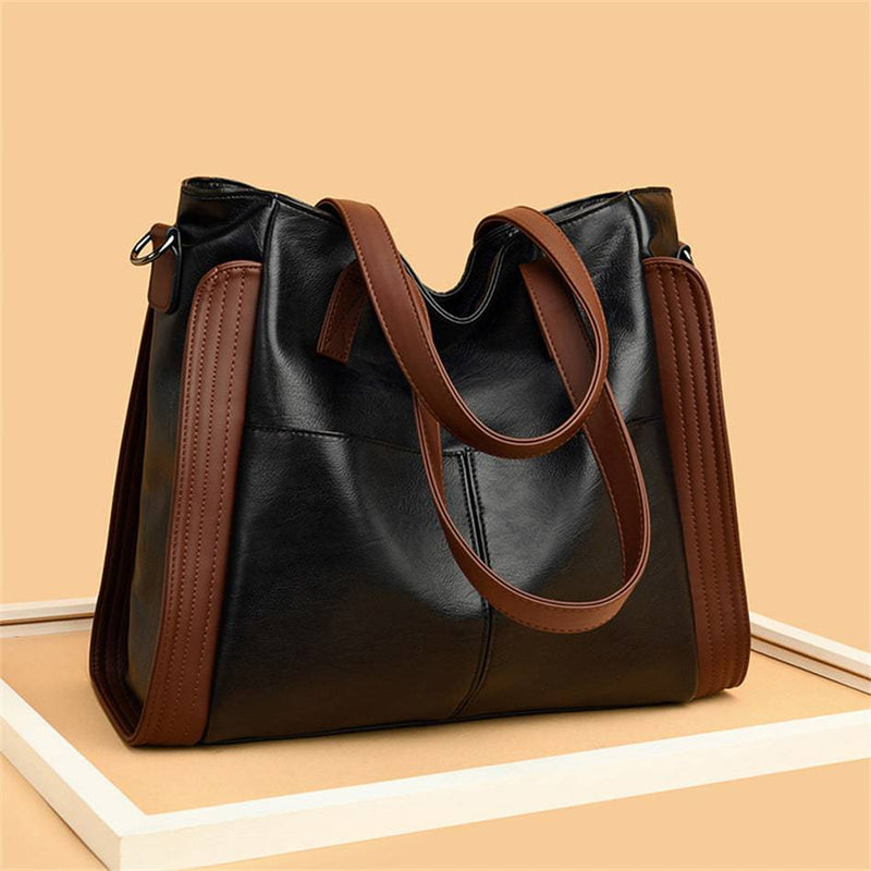 Bolsa tote feminina de couro sintético Sofia preta com detalhes marrons nas laterais e alças, exibida sobre uma plataforma branca em fundo bege, destacando seu design.