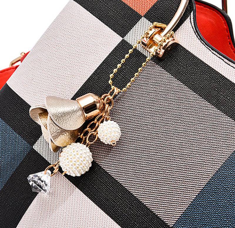 Detalhe em close-up do chaveiro da bolsa tote estampada, apresentando uma flor dourada, bolinhas peroladas e um cristal, pendurados por corrente ao lado da alça, em fundo xadrez.