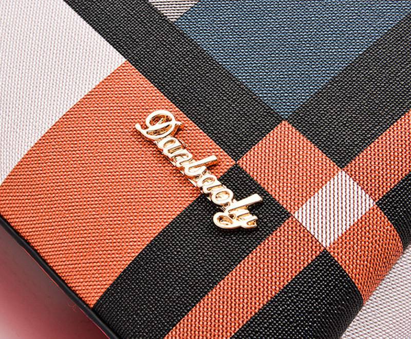 Detalhe em close-up do logotipo metálico dourado "Dualbeauty" na bolsa tote estampada, evidenciando o padrão xadrez multicolorido em tons de laranja, cinza e preto.