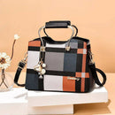 Bolsa tote feminina estruturada com estampa xadrez multicolorida em tons de cinza, laranja e preto, alça de mão de metal com detalhes em couro preto, e chaveiro de abelha.
