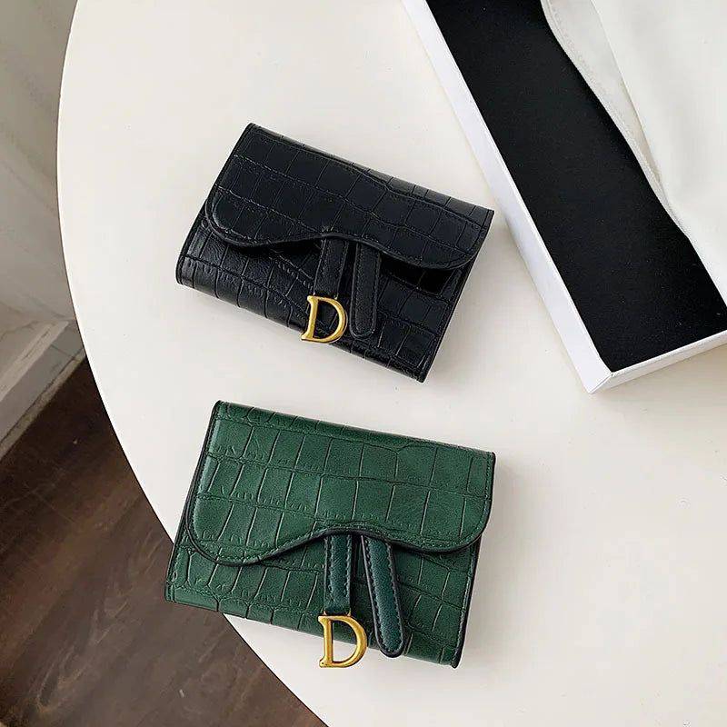 Duas carteiras de couro pequenas com textura de crocodilo, uma preta e outra verde escura, ambas com detalhe metálico dourado em forma de 'D', dispostas sobre uma mesa branca.