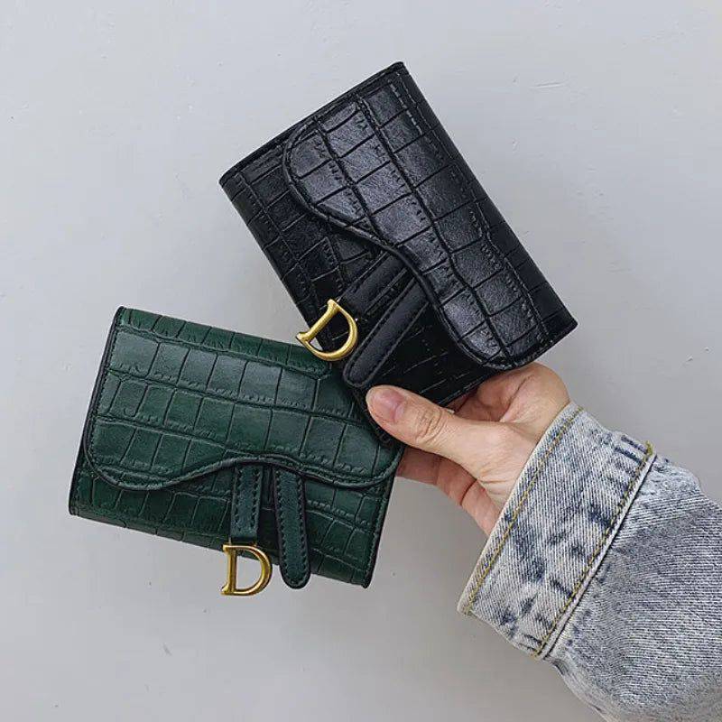 Mão feminina segurando duas carteiras de couro pequenas com textura de crocodilo, uma preta e outra verde escura, ambas com fecho de aba e detalhe metálico dourado em forma de 'D'.
