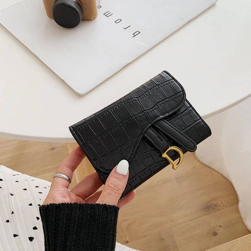 Mão feminina segurando carteira de couro pequena preta com textura de crocodilo e detalhe metálico dourado em forma de 'D', sobre uma mesa branca com um livro.