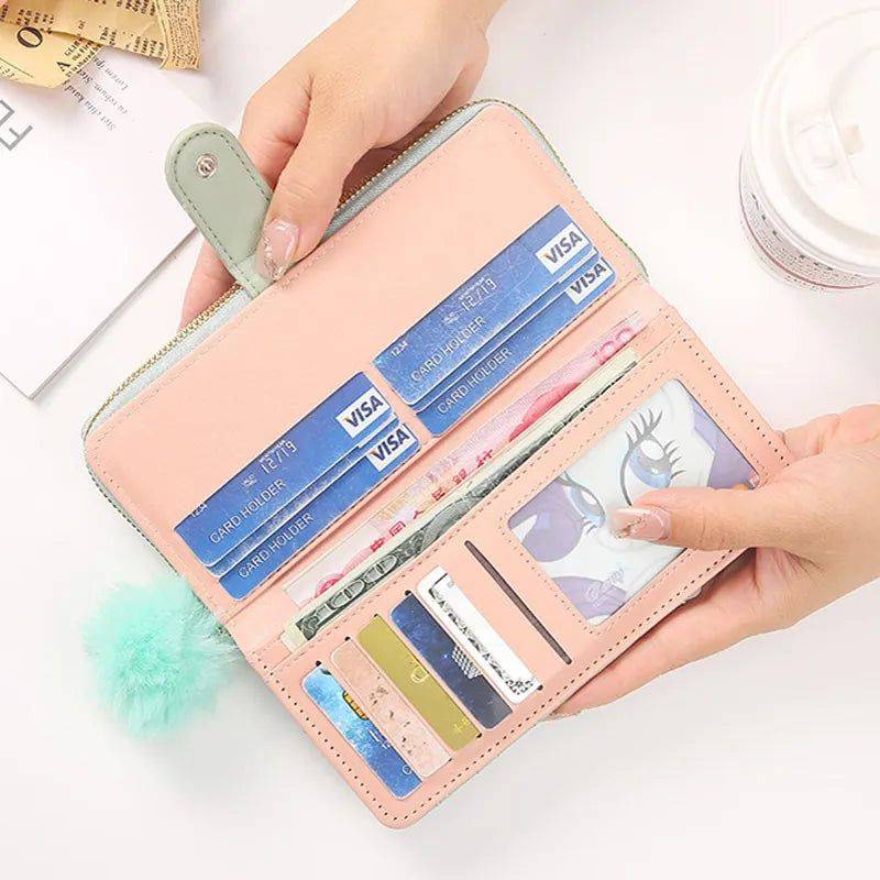 ãos femininas abrindo carteira de couro Aisha verde-claro, revelando slots para cartões, dinheiro e fotos, com acabamento em rosa no interior e pompom visível.