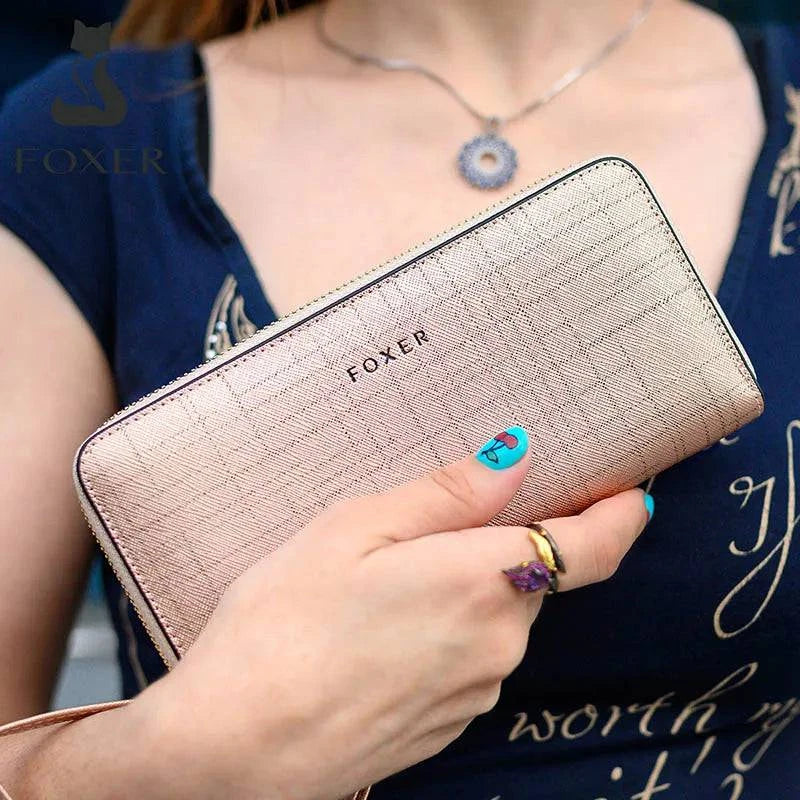 Mão feminina com anel e unhas azuis segurando carteira de couro Mônaco dourada/champanhe com textura quadriculada e logotipo "FOXER", combinando com colar e blusa azul.