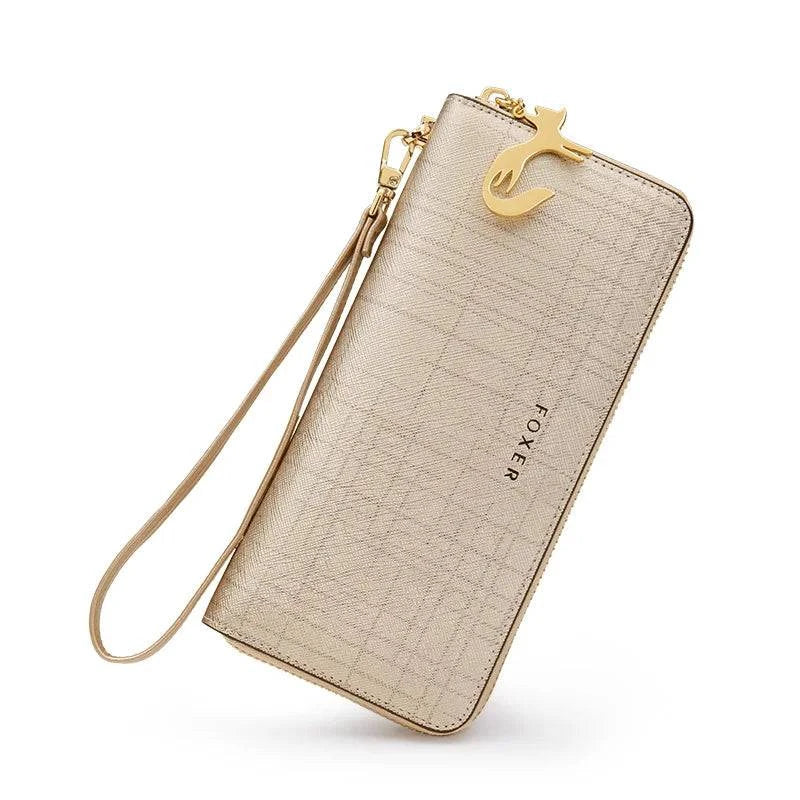 Carteira feminina de couro Mônaco dourada/champanhe com textura quadriculada, alça de pulso removível e charm de gato dourado, exibida em ângulo dinâmico sobre fundo branco.