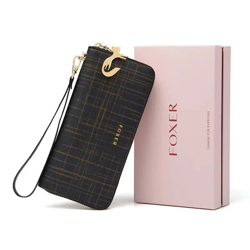 Carteira feminina de couro Mônaco preta com padrão quadriculado sutil, alça de pulso removível e charm de gato dourado, apresentada ao lado de sua caixa de presente rosa da marca FOXER.