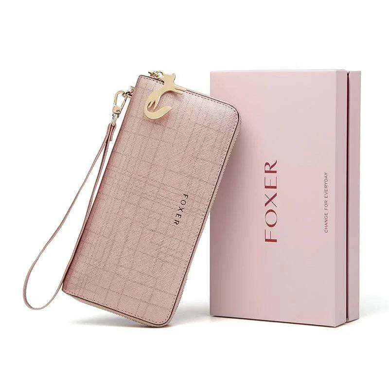 Carteira feminina de couro Mônaco rosa metálico com textura sutil, alça de pulso e charm de gato dourado, apresentada inclinada ao lado de sua caixa de presente rosa da marca "FOXER", ideal para presentear.
