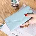 Mão feminina com unhas vermelhas segurando carteira de couro multifuncional azul-claro com fecho de aba e borla, sobre um caderno, com um copo descartável ao fundo.