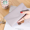 Mão feminina com anel dourado segurando carteira de couro multifuncional cinza com fecho de aba e borla, sobre um caderno, com um copo descartável ao fundo.
