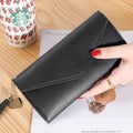 Mão feminina com unhas vermelhas segurando carteira de couro multifuncional preta com fecho de aba e borla, sobre um documento, com copo de café descartável ao fundo, em cena casual.