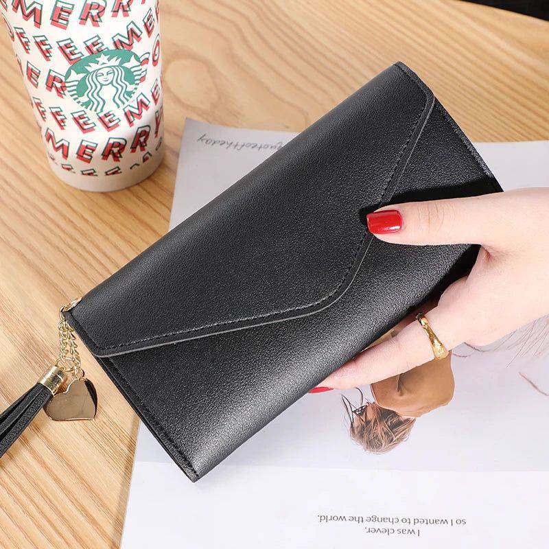 Mão feminina com anel dourado segurando carteira de couro multifuncional preta com fecho de aba, borla e chaveiro de coração, sobre um caderno, com um copo descartável ao fundo.