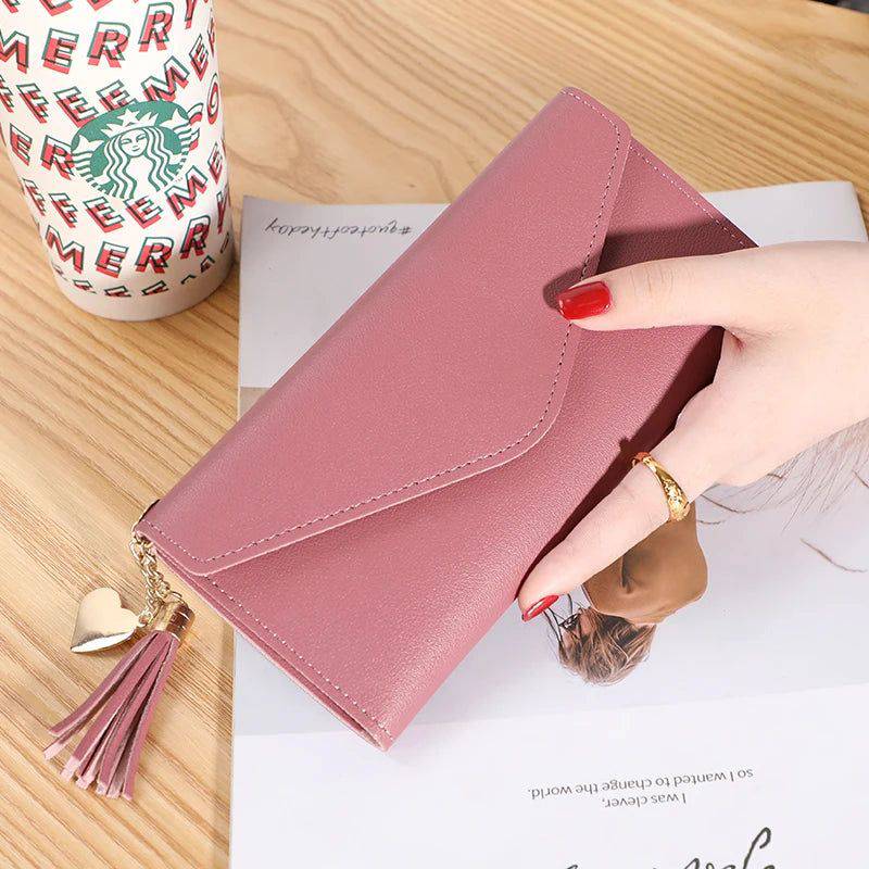 Mão feminina com anel dourado segurando carteira de couro multifuncional rosa com fecho de aba e borlas, sobre um caderno, com um copo descartável ao fundo, em cena casual.
