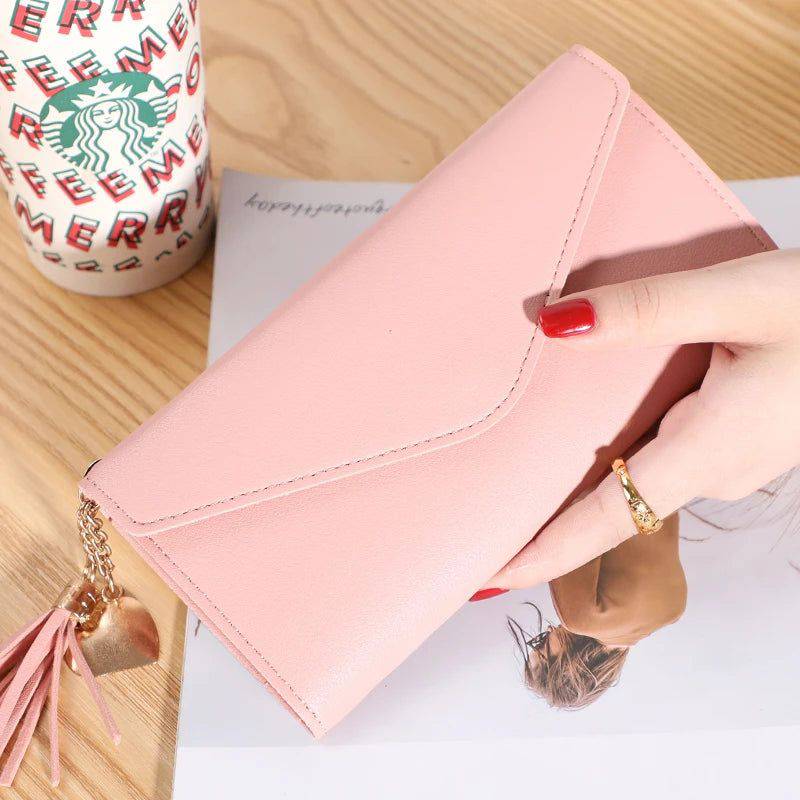 Mão feminina com anel dourado segurando carteira de couro multifuncional rosa claro com fecho de aba e borla, sobre um documento, com copo de café descartável ao fundo, em cenário casual.