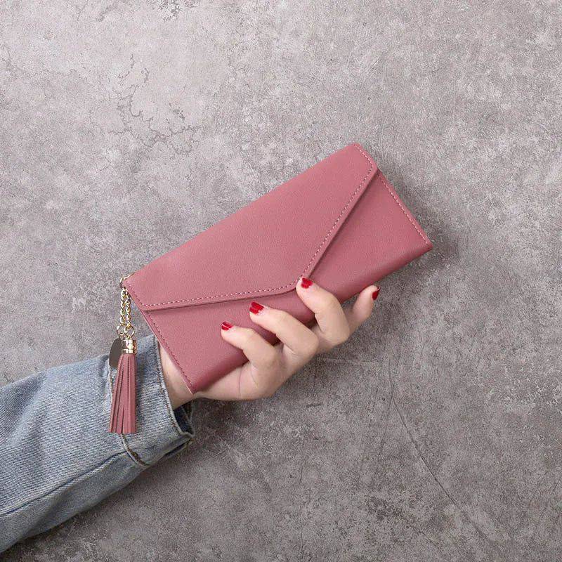 Mão feminina com unhas vermelhas segurando carteira de couro multifuncional rosa com fecho de aba e borla, contra uma parede de concreto, em estilo casual.