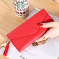 Mão feminina com anel dourado segurando carteira de couro multifuncional vermelha com fecho de aba e borla, sobre um caderno, com um copo descartável ao fundo.