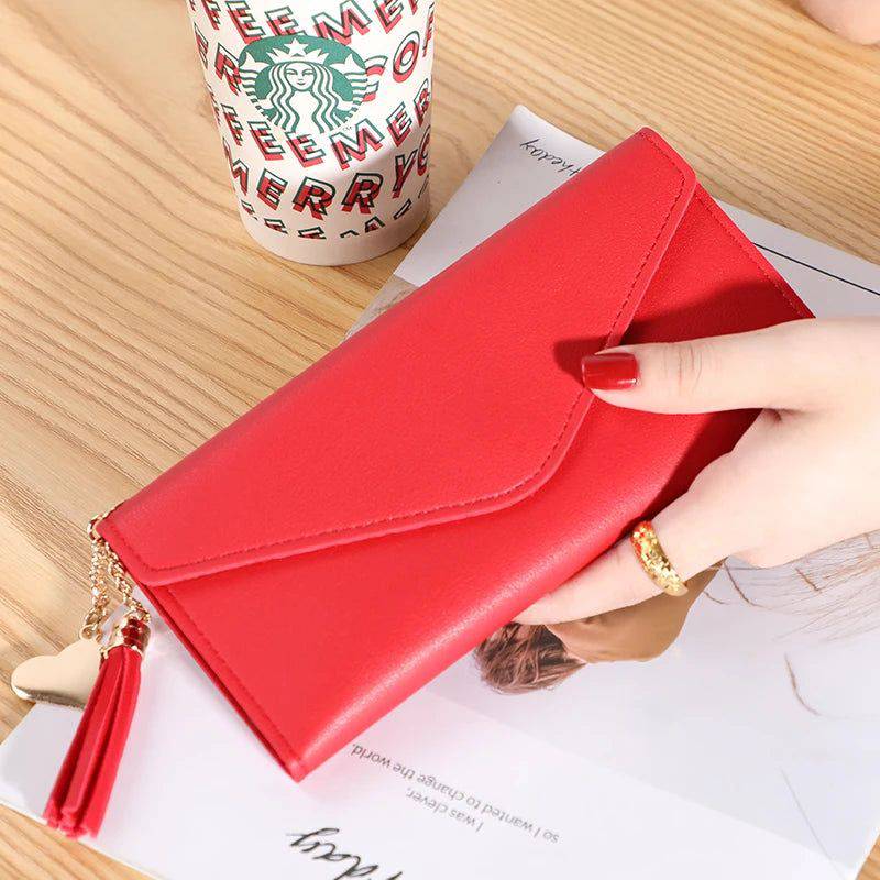 Mão feminina com anel dourado segurando carteira de couro multifuncional vermelha com fecho de aba e borla, sobre um caderno, com um copo descartável ao fundo.