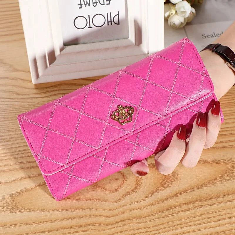 Mão feminina com unhas vermelhas segurando carteira feminina de couro sofisticada pink vibrante, acolchoada em matelassê com fecho de coroa dourada, sobre mesa de madeira.