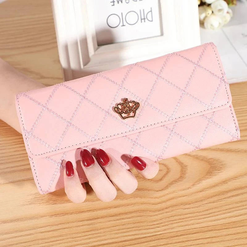 Mão feminina com unhas vermelhas segurando carteira de couro sofisticada rosa, acolchoada com padrão de diamante e fecho de coroa dourada, em mesa de madeira com porta-retrato ao fundo.