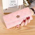 Mão feminina exibindo carteira de couro sofisticada rosa com design acolchoado de losangos e fecho de coroa dourada, sobre superfície de madeira clara com porta-retrato.