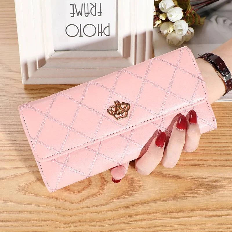Mão feminina exibindo carteira de couro sofisticada rosa com design acolchoado de losangos e fecho de coroa dourada, sobre superfície de madeira clara com porta-retrato.
