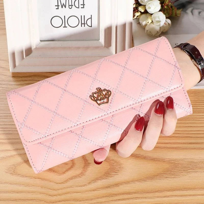 Mão feminina com unhas vermelhas segurando carteira feminina de couro sofisticada rosa, acolchoada em matelassê com fecho de coroa dourada, em ambiente interno.