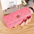 Mão feminina com unhas vermelhas segurando carteira de couro sofisticada rosa vibrante, com design acolchoado em losangos e fecho de coroa dourada, sobre mesa de madeira.