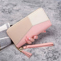 Mão feminina com unhas peroladas segurando carteira de couro com zíper e alça, em bloco de cores rosa, bege e branco, com alça de pulso rosa claro e borlas, sobre fundo de concreto.