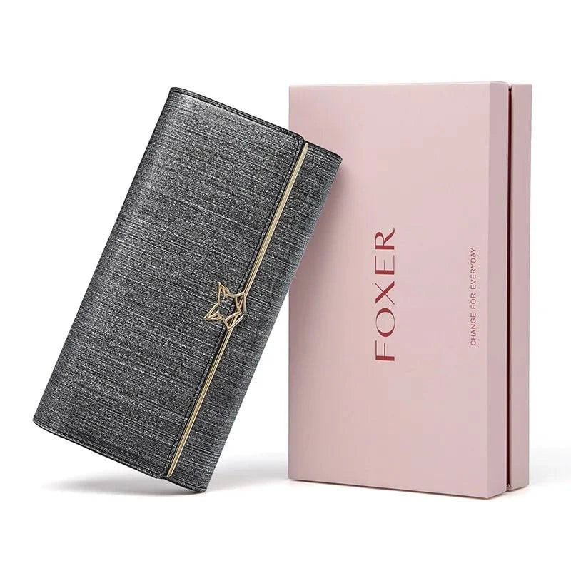 Carteira feminina de couro luxuosa cinza metálico com textura sutil e fecho de estrela dourado, exibida inclinada ao lado de sua caixa de presente rosa da marca "FOXER".