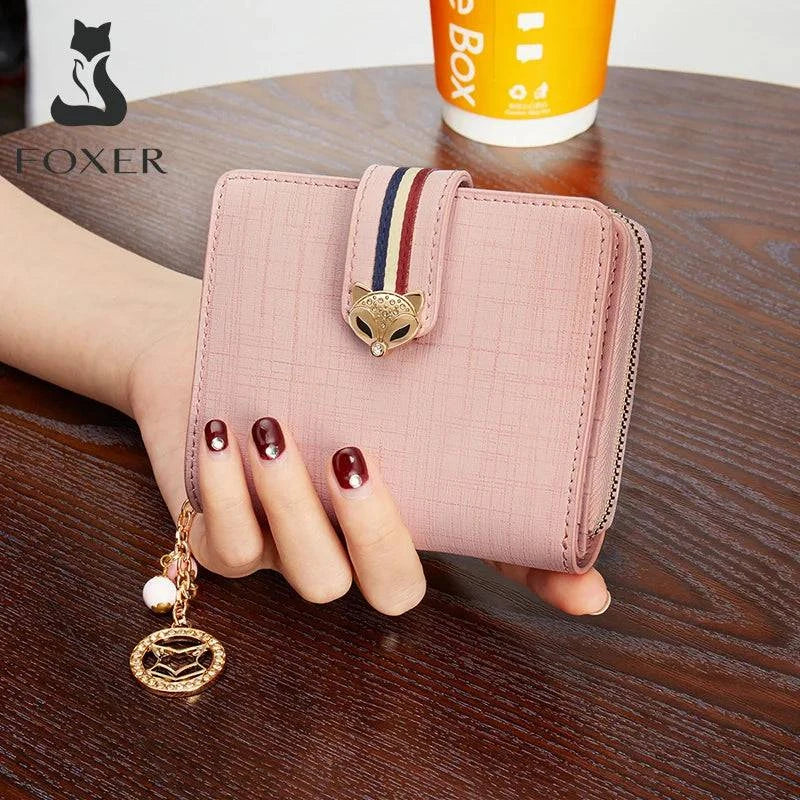 Mão segurando carteira feminina pequena elegante de couro rosa claro com fecho de raposa dourado e tira listrada, exibida sobre mesa de madeira, com logotipo FOXER.