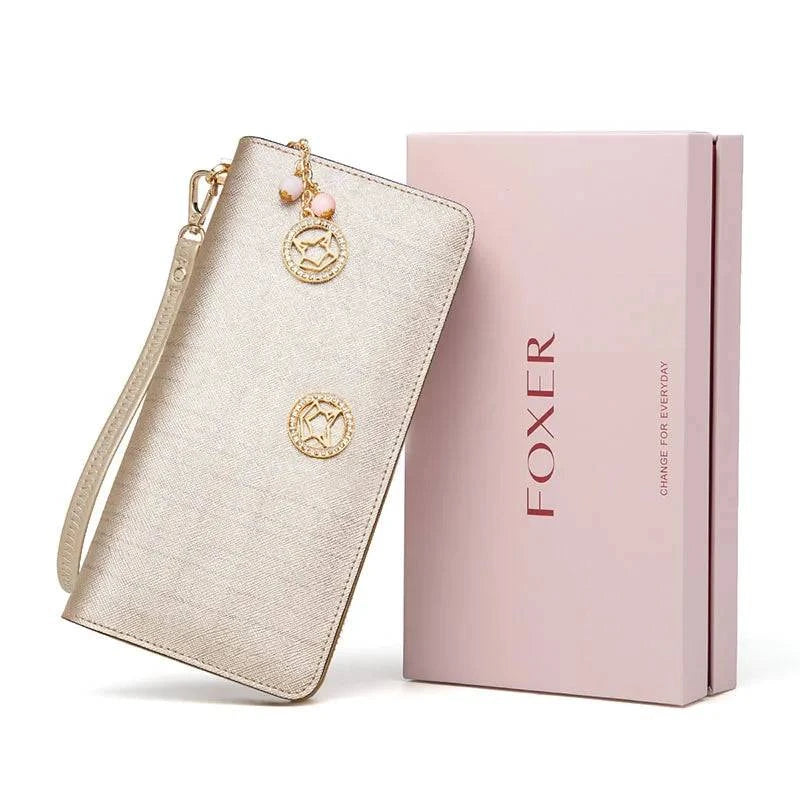 Carteira feminina de couro premium dourada/champanhe com textura fina e medalhão de raposa, exibida inclinada ao lado de sua caixa de presente rosa da marca "FOXER", ideal para presente.