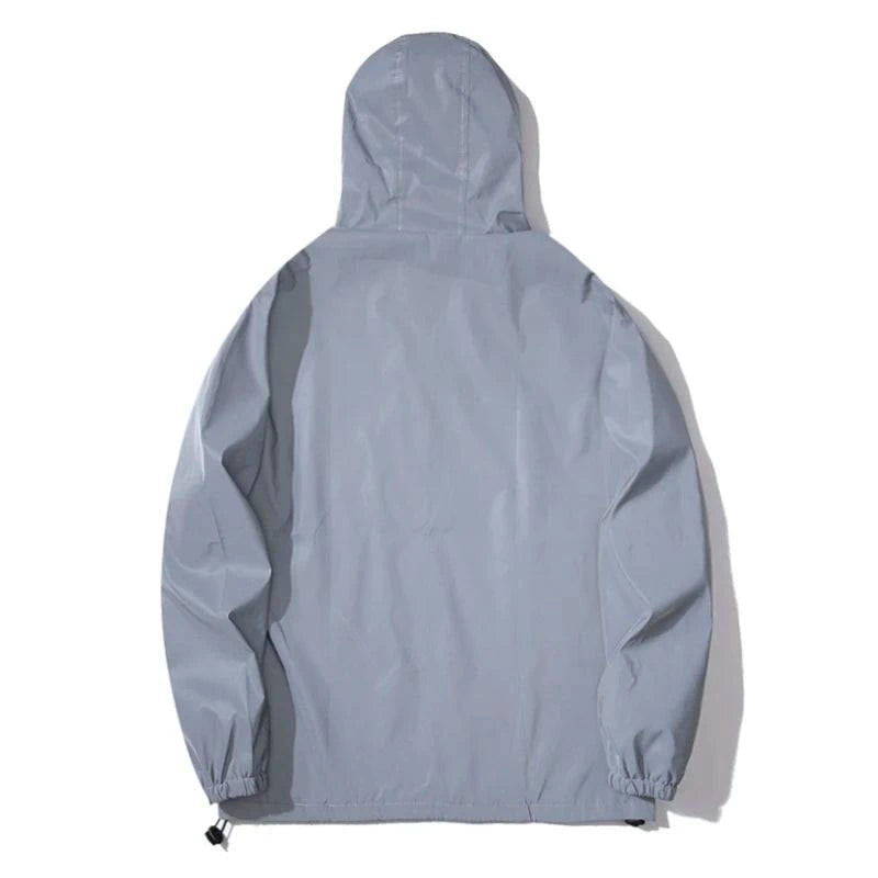 Jaqueta corta-vento em tecido totalmente refletivo, aparece em um branco brilhante contra um fundo preto, destacando o capuz e os zíperes pretos nos bolsos e frontal. Design de alta visibilidade e exclusividade para segurança noturna.