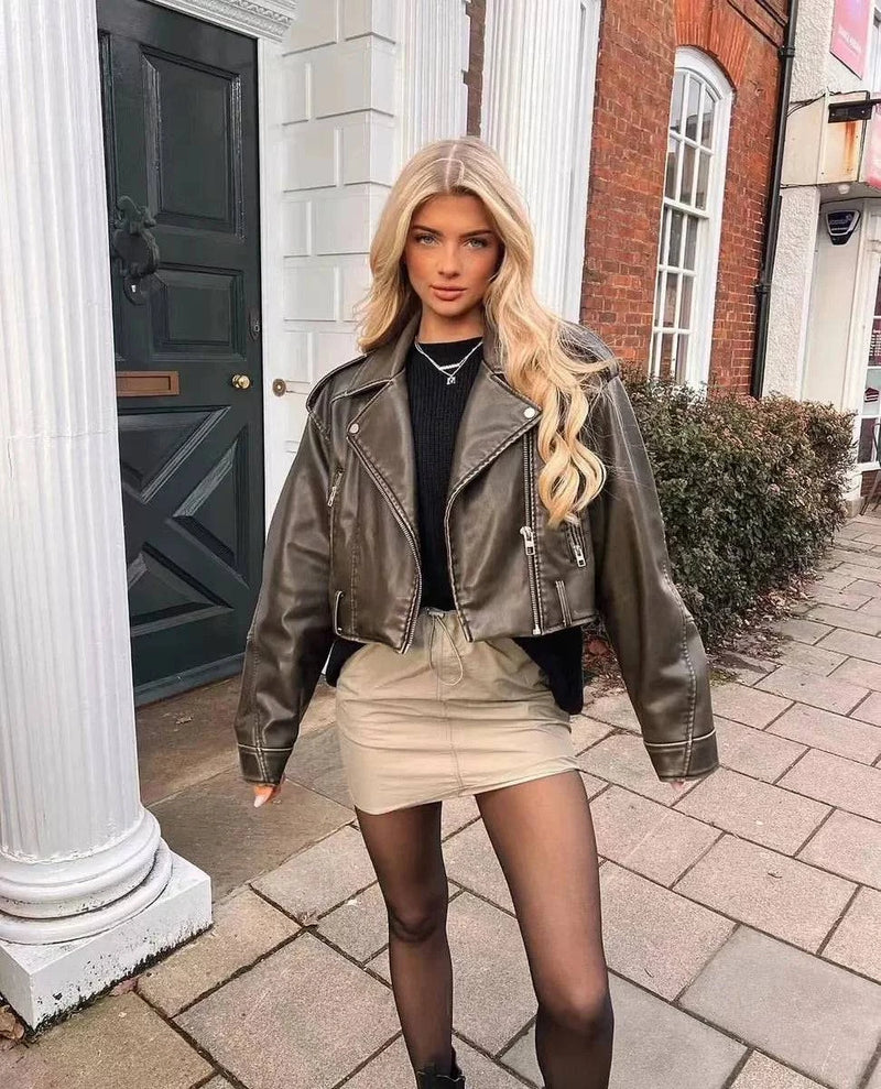 Mulher loira usando jaqueta de couro feminina marrom com acabamento distressed, combinada com saia bege e meia-calça preta, criando um visual urbano e sofisticado de alto padrão.