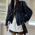 Mulher vestindo jaqueta de couro feminina preta, estilo biker com zíperes e cinto, combinada com camiseta branca e shorts, exibindo um visual urbano de luxo e atitude atemporal.